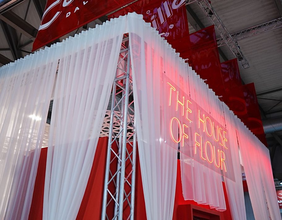Lo stand The House of Flour in fiera, con tende bianche e insegna al neon.